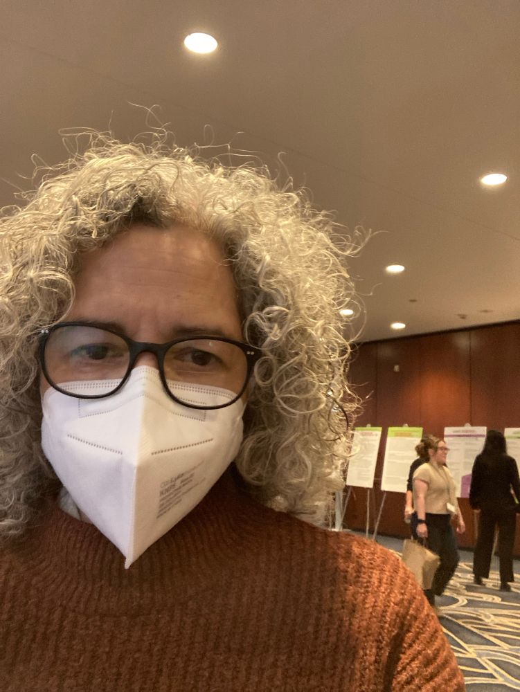 Adria Quinones at the Hilton : she has a lot pf gray hair, glasses and is wearing a mask. Over her left shoulder is a wood panelled wall; the conference schedule is posted on a series of tripods in front. A few women are reading and passing by. 