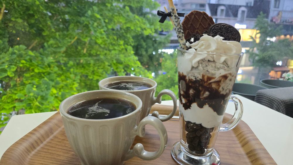 A tall oreo parfait and two cups of americanos sit on a tray. Behind it is a window that is revealing some rather poor weather...
