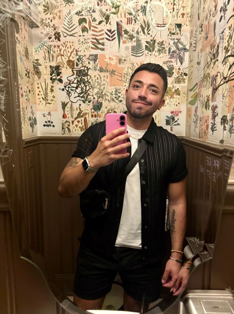 selfie of me in a bathroom wearing a black buttoned up and a white tank top showing my pink iphone 16