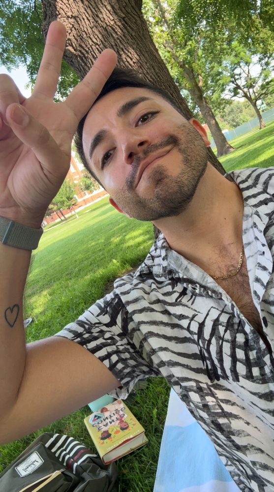 selfie of me at the park under a tree. i’m wearing a black and white stripped buttoned up and gold chain holding up a peace sign, sitting on a beach towel 