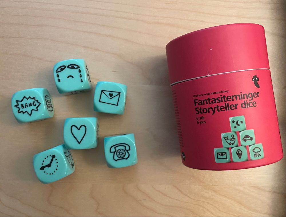 A picture of a red tin that says storyteller dice in English and in dutch. It shows six teal die that each to pick something different, in this case 1 says bang, another has a sad crying face, 1 has an envelope, 1 has a heart, 1 has a telephone and one has a clock