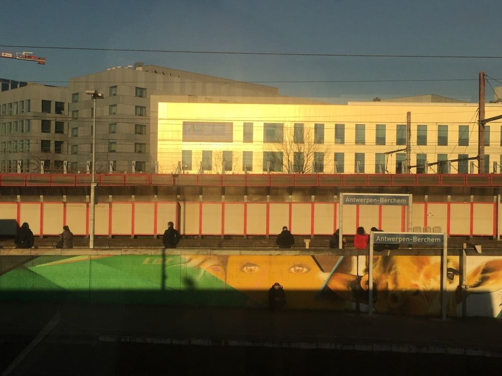 Passing through Antwerpen-Berchem station on a train. In the background, beneath an oppressively blue sky: a concrete building lit up by sunlight, some people waiting on a platform, and part of a mural. 