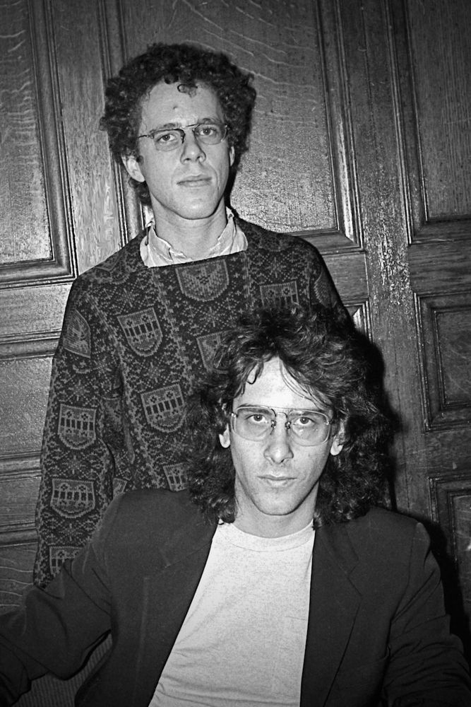 Ethan Coen is wearing a fancy jumper and standing behind his brother Joel Coen, who has longer hair and is wearing a smartish jacket over a t-shirt. In the background is wood panelling.