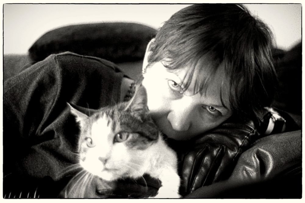 Black and white close-up of me, looking through my fringe at the camera and hugging a tabby & white cat who has spotted something more interesting on the other side of the room.