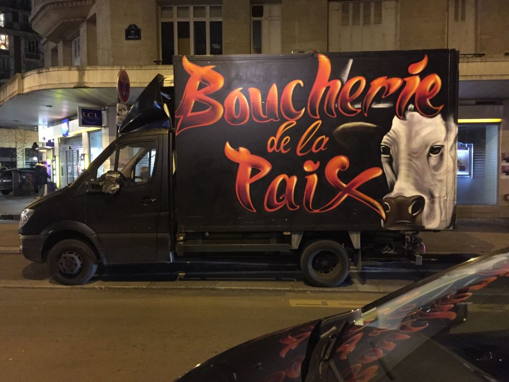 Paris by night: a black truck with "Boucherie de la Paix" written on the side in flamboyant 3D-effect red lettering next to a picture of a cow's face looking lugubrious. The red lettering is reflected in a parked car in the foreground. It's like a scene out of some kind of Vegetarian Hell.