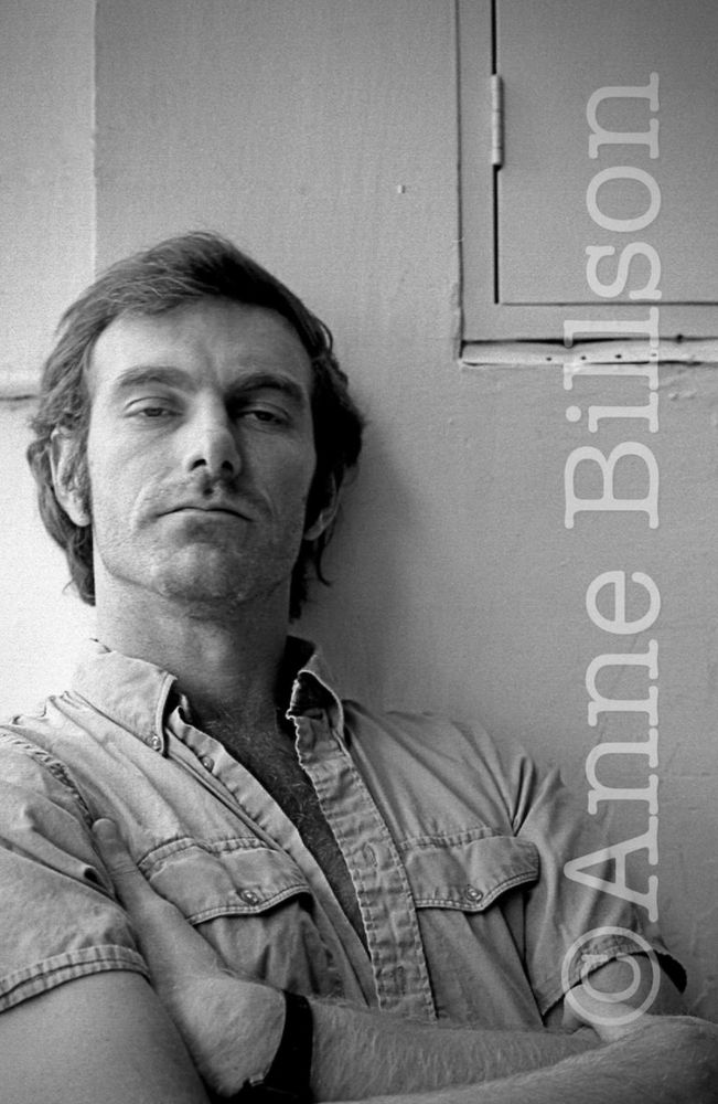 Black and white photograph of the filmmaker John Sayles, arms crossed in a short-sleeved denim shirt.