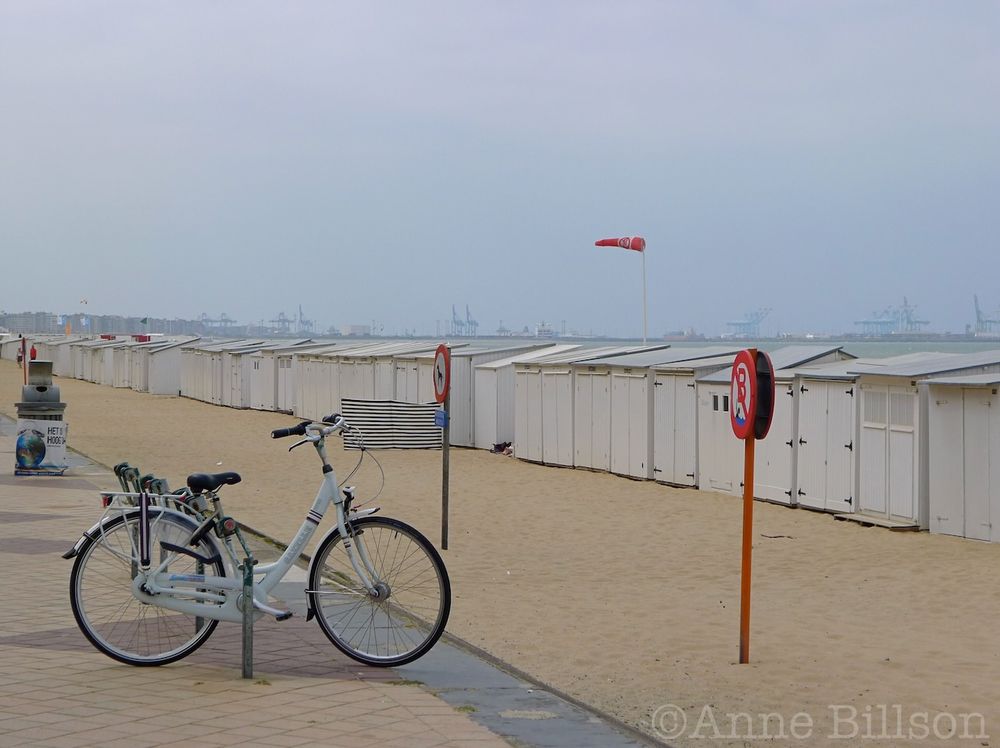 A parked bicycle and a row of beach huts on a seafront that seems somewhat drained of colour, apart from an orange wind sock (fully employed) and a no-bathing sign. The sky is grey. On the distant horizon can be glimpsed faint outlines of the port of Zeebrugge's massive industrial cranes.