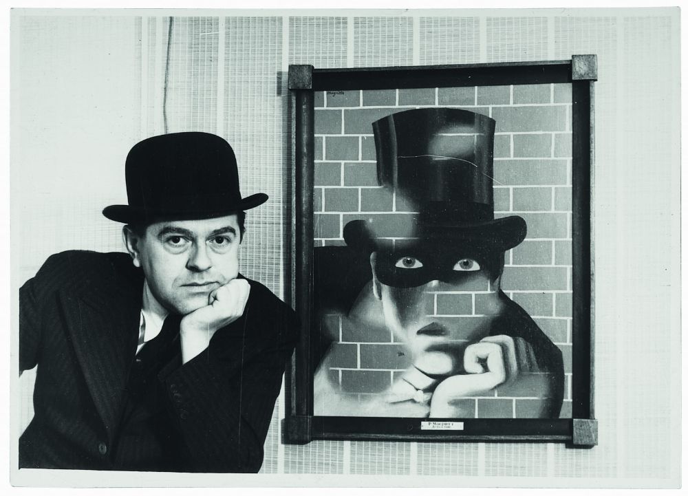 Magritte, in his bowler hat, echoes the chin-on-hand pose of the tophatted, masked antihero Fantômas, whom he has painted looming out of a brick wall.