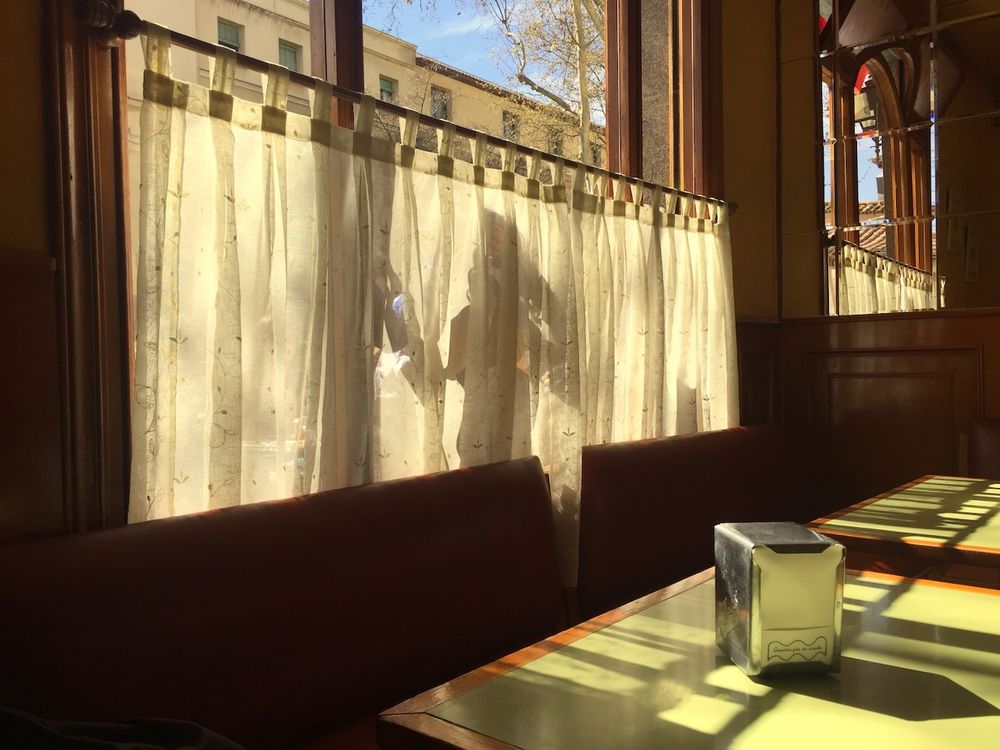 The interior of an apparently deserted café; the sun is shining through the window, casting the shadow of a woman on her phone on the net curtaining, and, inside, geometric shapes over the green formica-topped tables, on one of which is standing a silver napkin dispenser. 