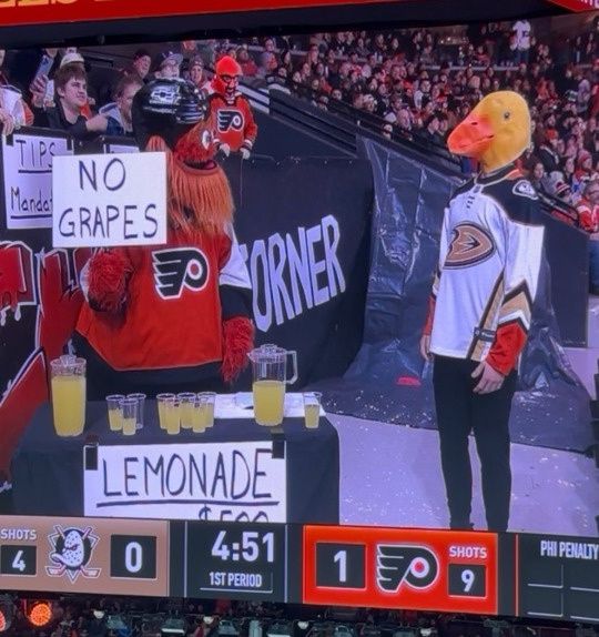 It’s a shot from a hockey match between the Philadelphia Flyers and the Anaheim Ducks, but no hockey is actually seen (though the score is 0-1 Flyers, with 4:51 left in the first period). Instead, a corner of the arena is here. Gritty, the Flyers’ beloved eccentric, Mr. It themed mascot, has set up a lemonade stand in a corner of the arena, surrounded by fans. He is selling lemonade for an exorbitant price. He is also holding a sign up that says “No Grapes”. The Ducks’, well, duck mascot looks on with a look of dejection on its face. It’s a wonderful reference to the children’s song, The Duck Song and I keep laughing at it. I assume the duck waddles away sadly after this. 