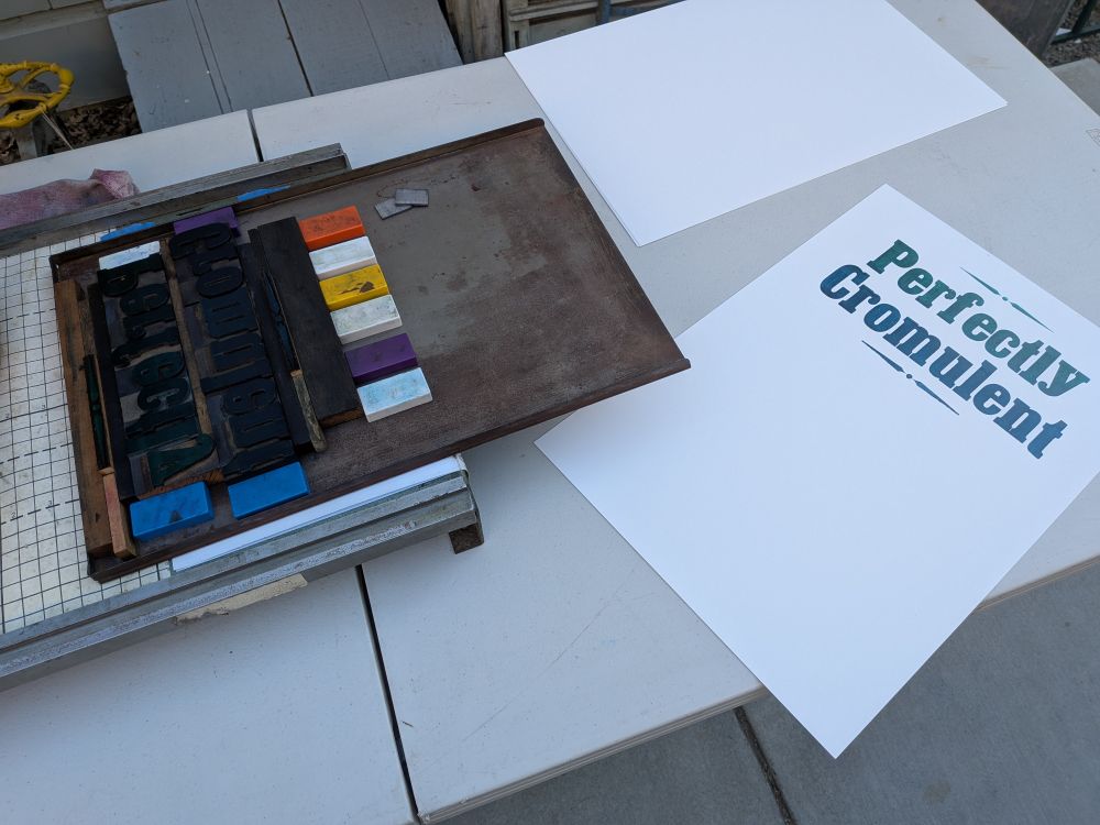 Metal galley tray with wood type held in place with heavy-duty magnets, sitting on top of a small poster press. The first finished print sits off to the side.