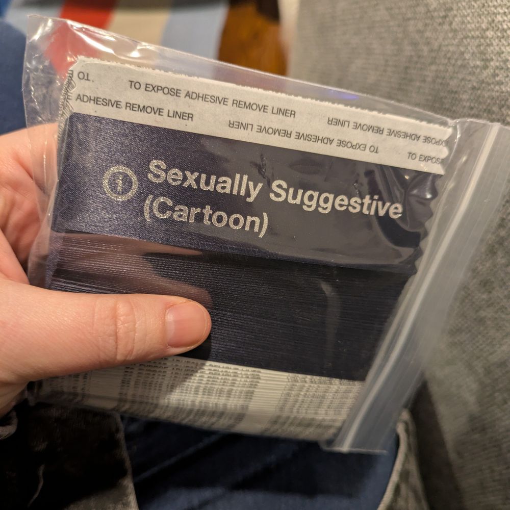 A navy blue badge ribbon with silver text reading: "Sexually Suggestive (Cartoon)" stylized like the Bluesky moderation tag