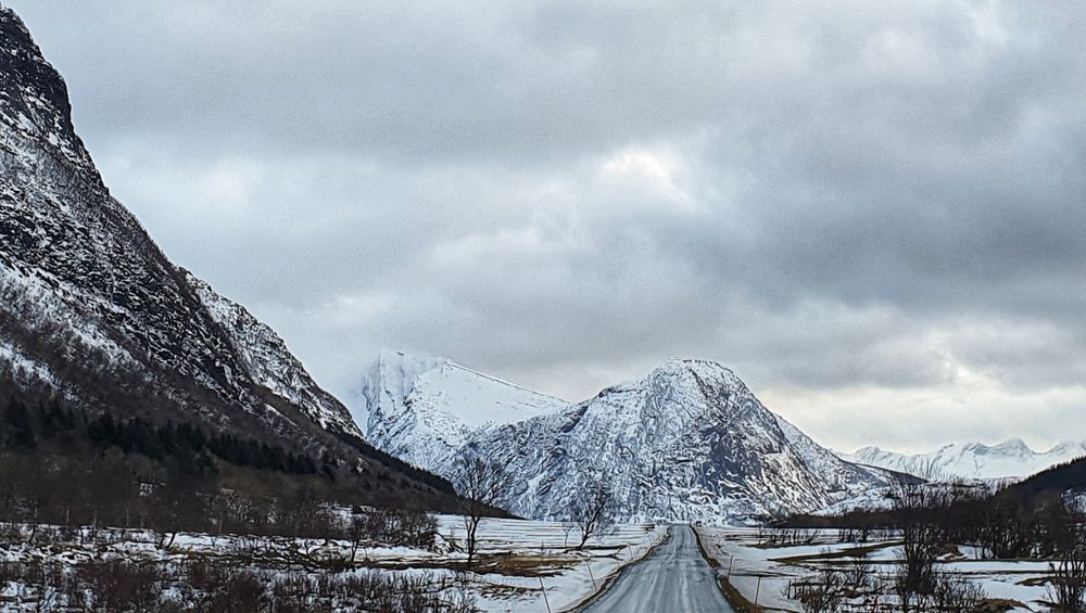 En grå, humpete asfaltvei mellom og foran høye, snøkledte fjell
