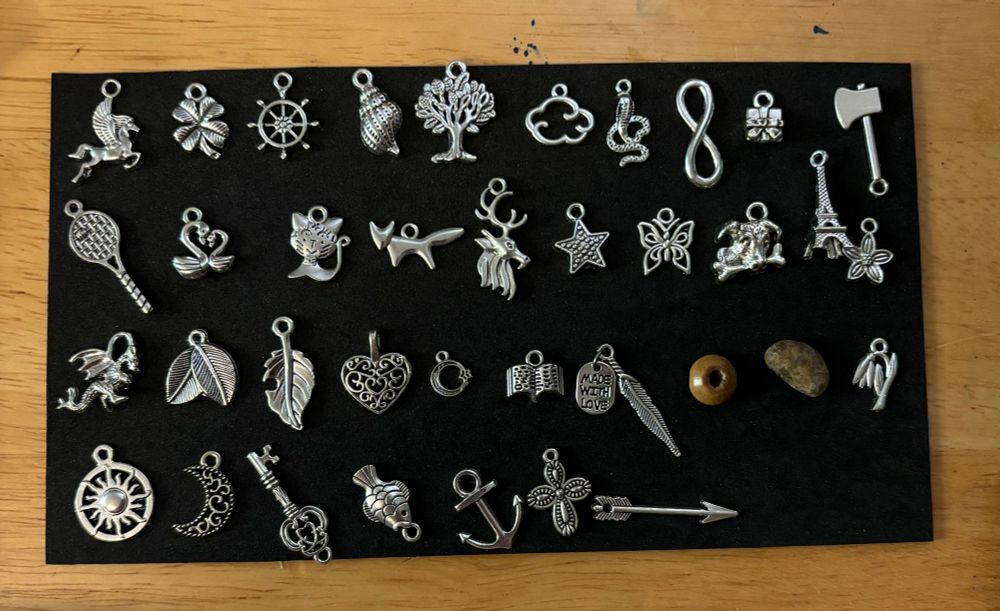 A set of silver charms laid out on a black foam background. The charms are in order of the Lenormand system with the addition of one, an arrow charm at the end. 