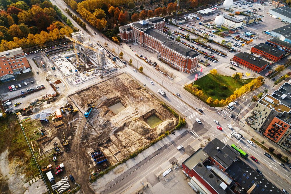 Drone image of a city. In the middle a building site. Around it streets, parking spaces and houses.