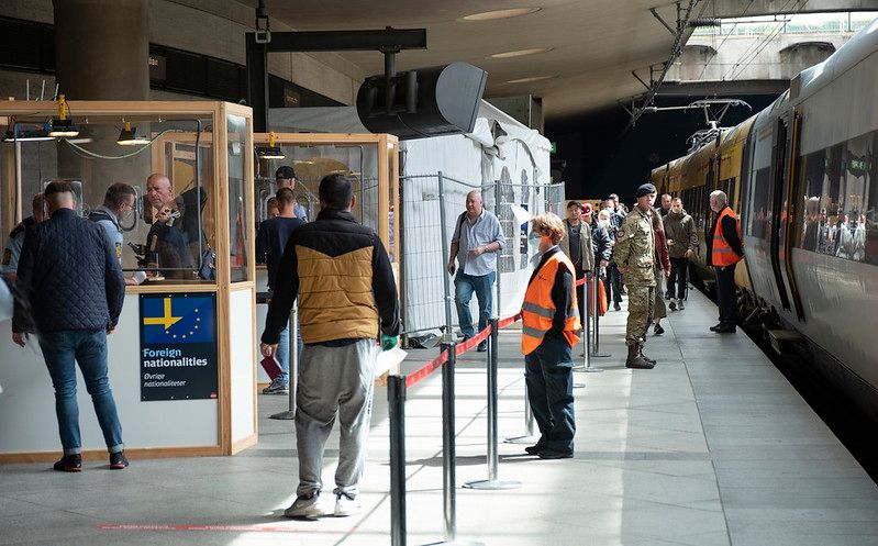 A photograph of people moving between a train, a fence and booths. A person in military uniform is standing close to the train.