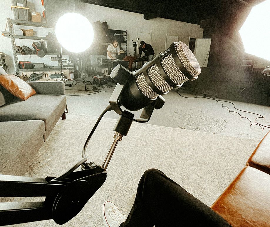 A photo of a microphone. Behind it a room with lights, sofas, and two people working at a desk.
