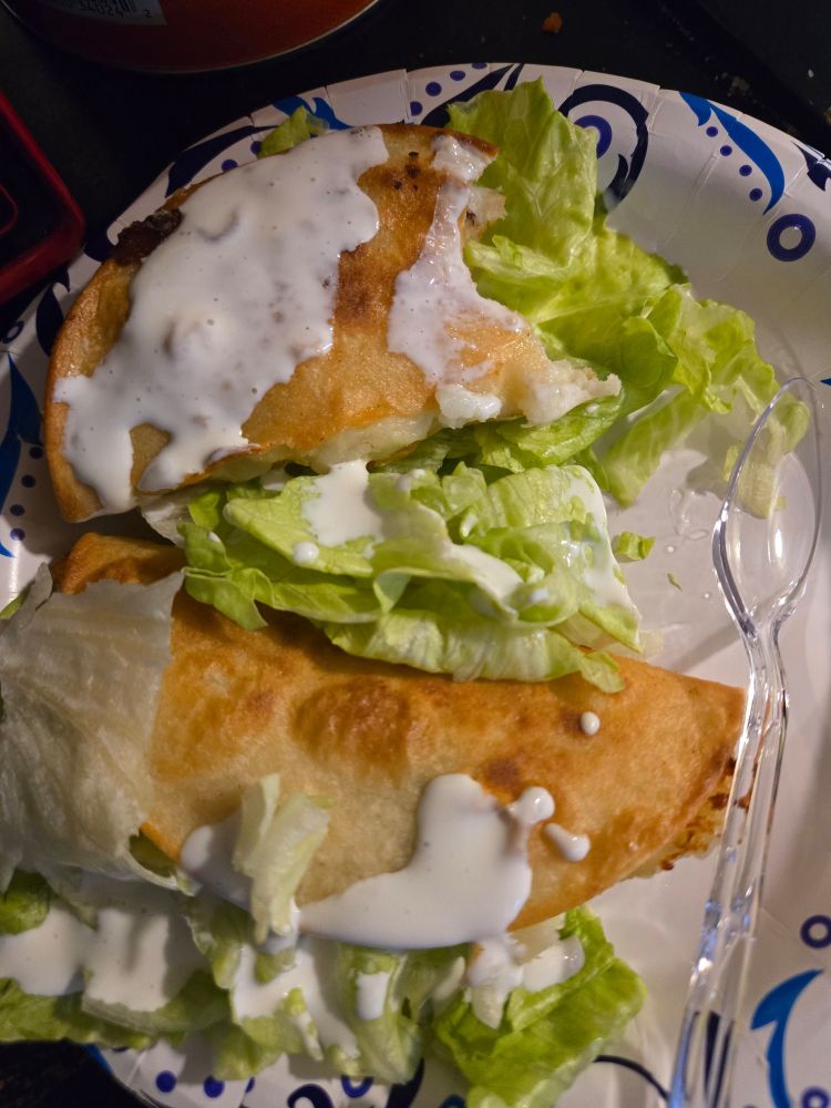 Two pan fried tacos de papa on a paper plate, stuffed with some hand shredded iceberg lettuce and drizzled with crema. A plastic spoon sits to the right