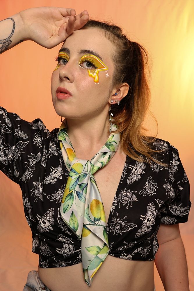 A young woman with light skin and a confident expression sits against a warm orange gradient background. She wears bold yellow and white eye makeup with intricate butterfly-like designs extending from her eyes. Her hair is styled in a high ponytail, with some strands framing her face. She has multiple ear piercings and a tattoo on her left forearm. She is dressed in a black cropped blouse with a white moth pattern, high-waisted light-wash jeans, and a lemon-patterned scarf loosely tied around her neck. Her pose is relaxed