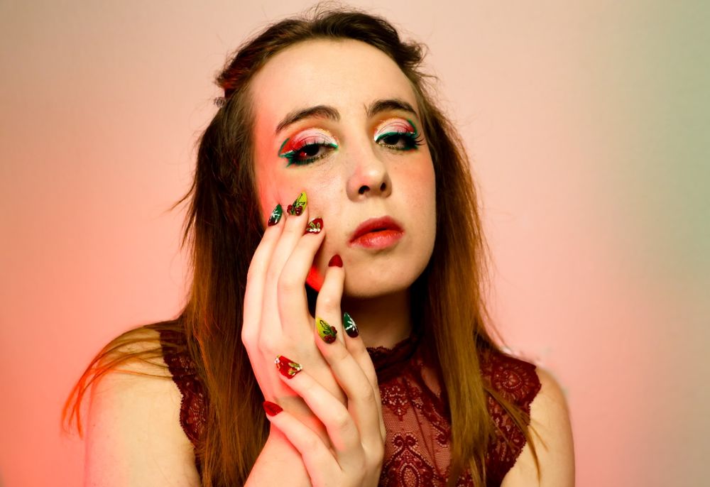 A person with long brown hair and red white and green makeup. Touching the right side of their cheek with their hand. They are wearing a red top 