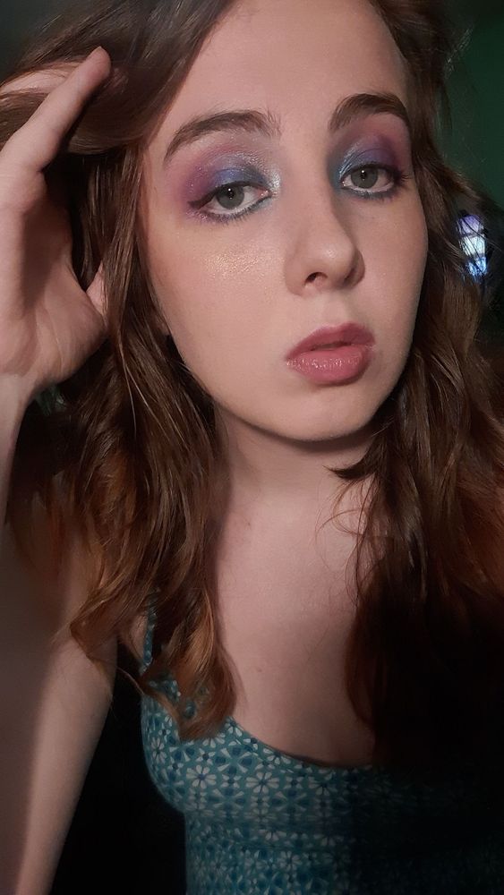 A person with brown hair. Their makeup is purple near the outer corners of the eye and blends into blue towards the inner corner of the eyes. They have a subtle wing and mascara and a pink lip. Their hand is in their hair and they are wearing a blue geometric pattern dress 