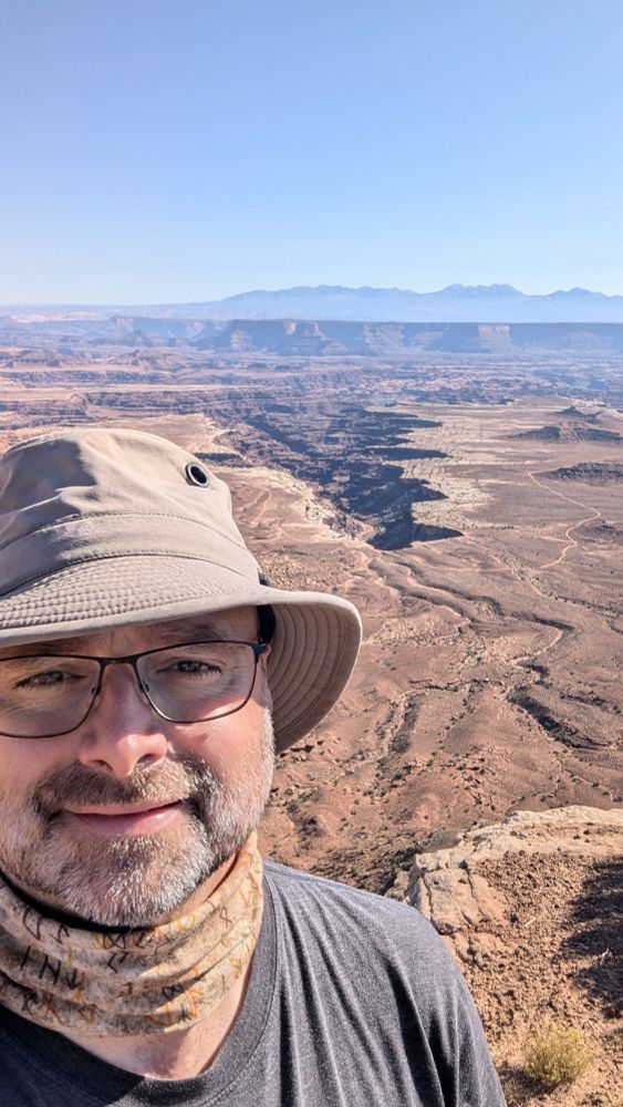 Man standing in front of a desert valley landscape. The breaded man has glasses, a grey Tilley hat and a brown bandana on. The landscape shows a distant river valley that has eroded through millions of years of sedimentary rock.