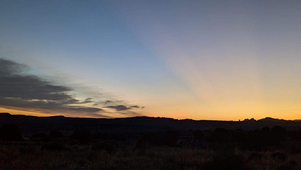 Dark landscape with the sun getting closer to popping over the far Hills. Crepuscular rays are showing from behind a far peak. Clouds have appeared on the left side.