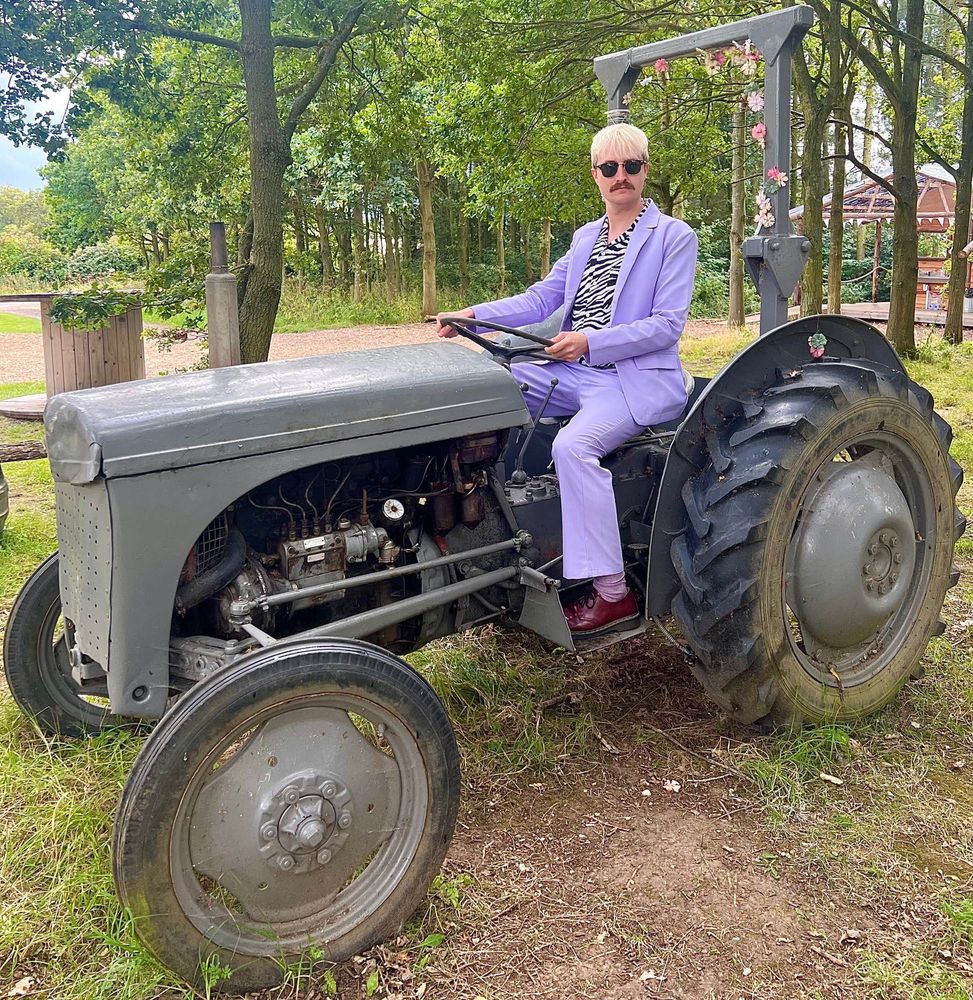 Picture of myself a white man with bleach blonde short hair & moustache wearing a pastel purple suit jacket & trousers with a black & white zebra print shirt riding a tractor in a field 