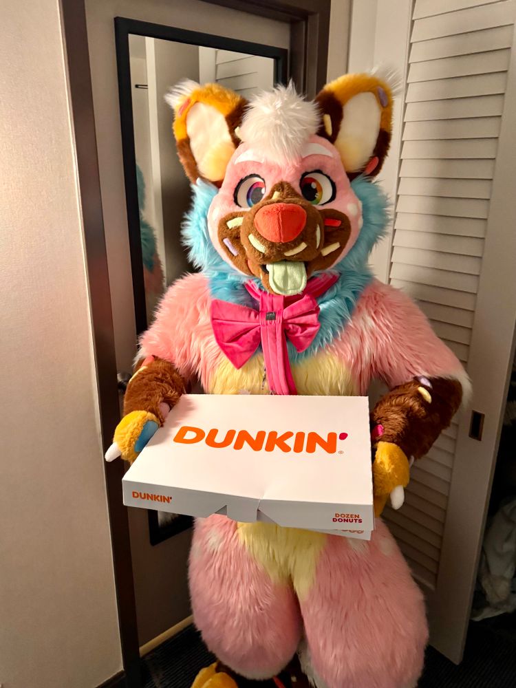 a pink sprinkledog holding a box of Dunkin’ Donuts! don’t actually take any because he secretly wants it all!
