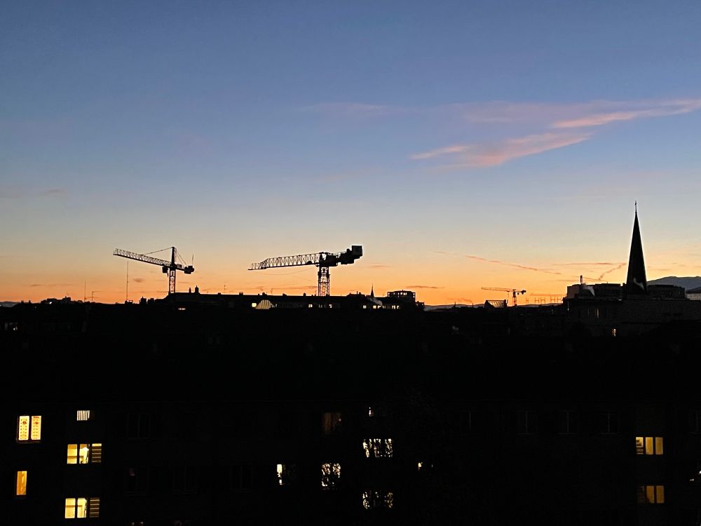 A sunset sky with orange near the horizon, up to a medium blue at the top of the frame. In the foreground are apartment buildings, with two construction cranes and a church spire towering over everything else. 