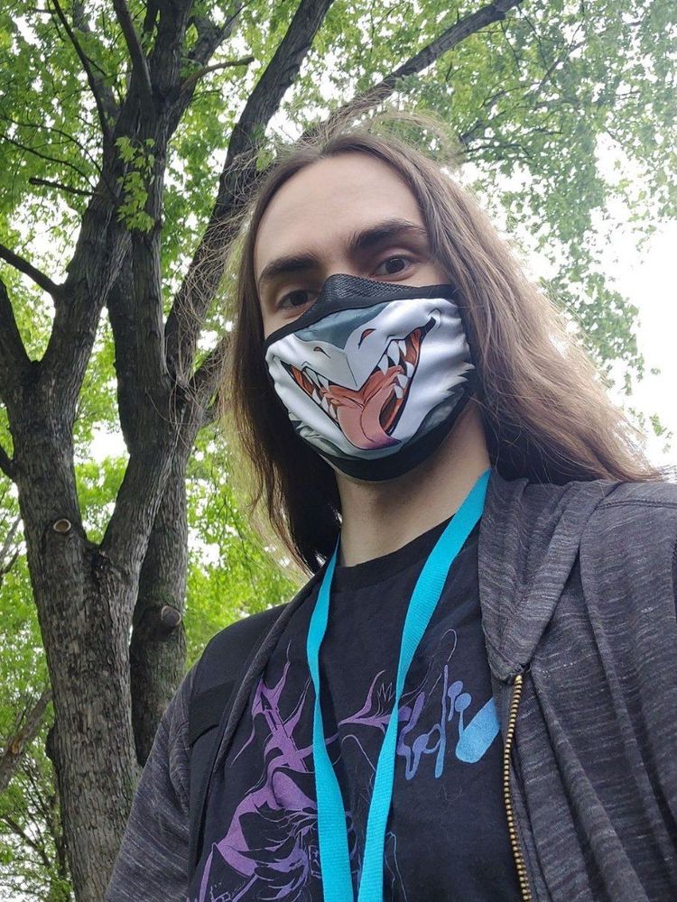 Selfie of cVoid in a sergal maw mask, teal/purple nevrean shirt, heathered grey hoodie, and teal lanyard in front of trees