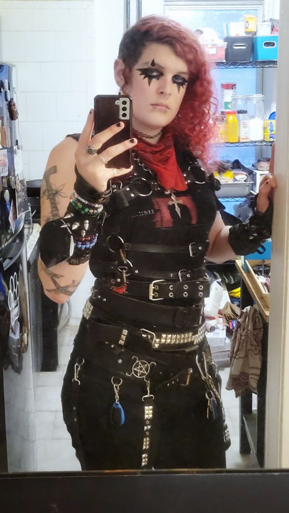 lilith (goth trans woman with red hair) taking a picture in a mirror. she is wearing many belts. she looks tall as fuck lol