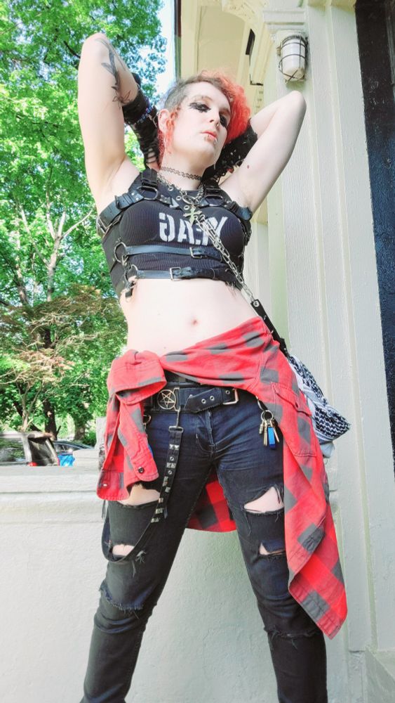 lilith (goth trans woman with red hair) posing with her arms up behind her head. she has a red flannel tied around her waist. her goth makeup is all fucked up. she looks fucking tired