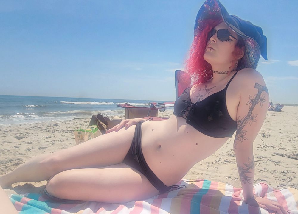 lilith (goth trans woman with red hair in a witch hat) on the beach in a black two piece swimsuit lounging on a beach towel. her waist looks dabonkers