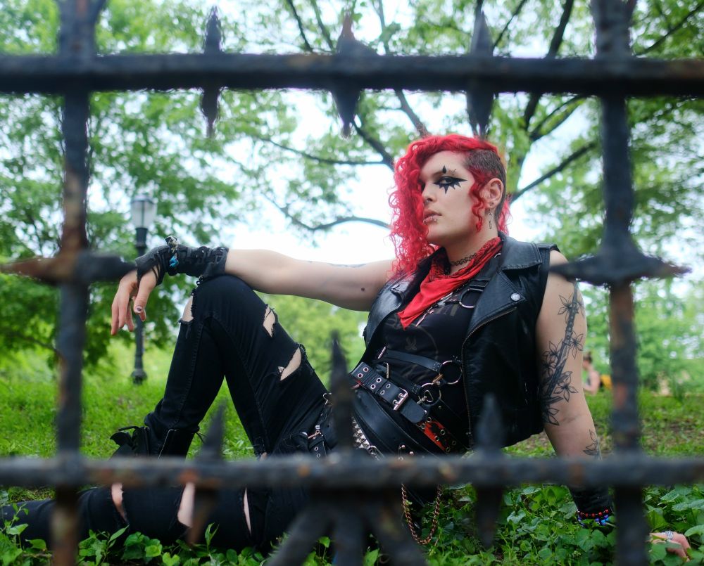lilith (goth trans woman with red hair) sitting on the ground with her right knee up and her right arm draped over it in front of a wroght iron fence in the foreground, out of focus