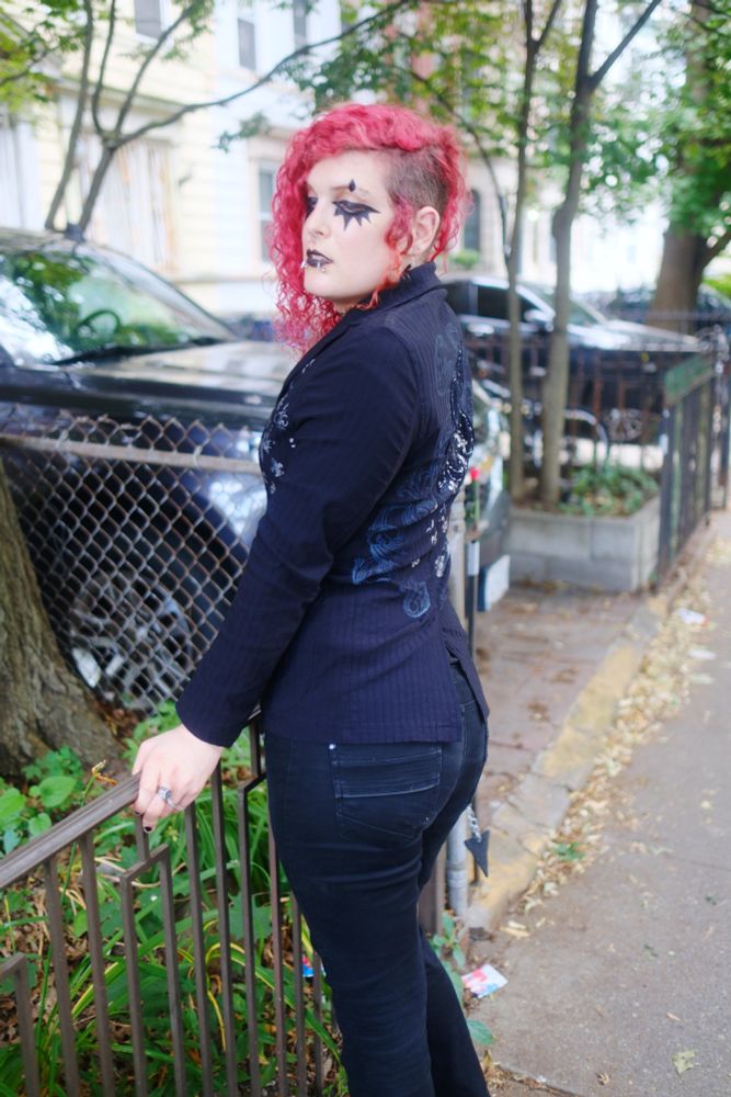 lilith (goth trans woman with red hair) wearing black pants and a blazer facing away from the camera. her butt looks very voluptuous