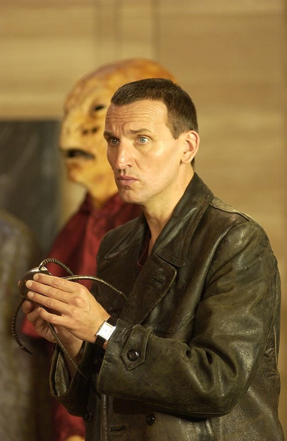 A screencap from Doctor Who and episode called The End of the World (2005) starring Christopher Eccleston as the 9th Doctor he is wearing his signature black leather jacket and holding one of the spiders Cassandra used to kill on board the station 