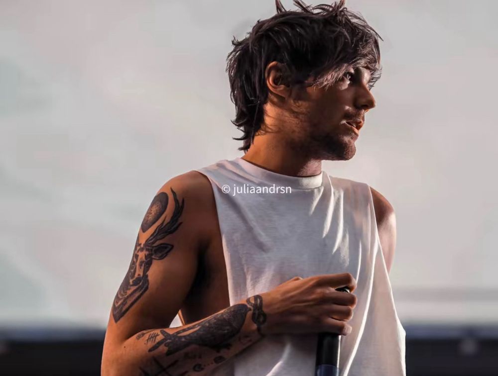 An unseen close-up of Louis Tomlinson on stage. He’s wearing a white shirt with the sleeves cut off, holding his mic to his chest, and his head is turned away. 
