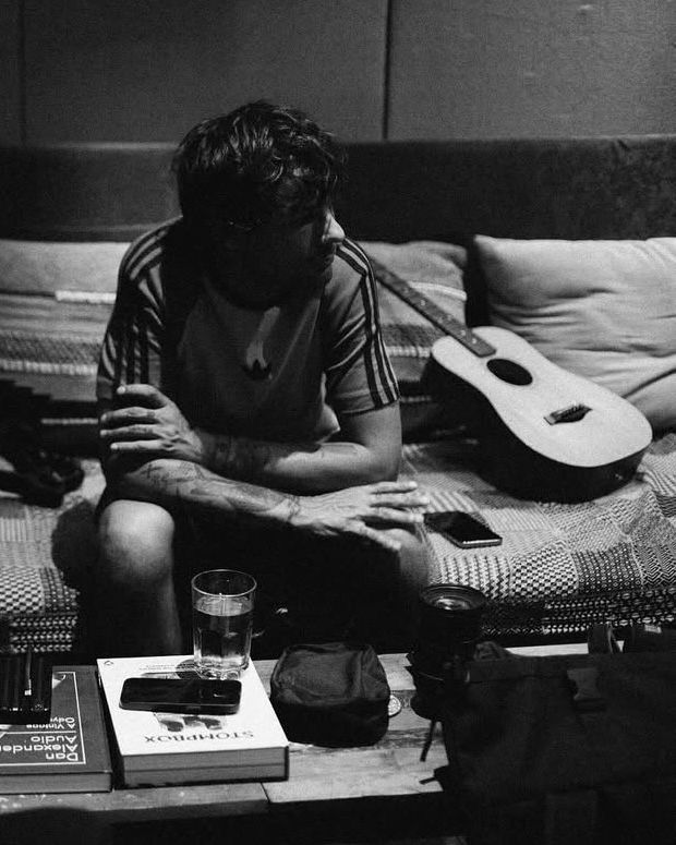 A cropped black and white photo of Louis Tomlinson sitting in the couch with his guitar. His arms are folded across his lap, his phone is next to him, and a glass of water is on a stack of books on the coffee table.