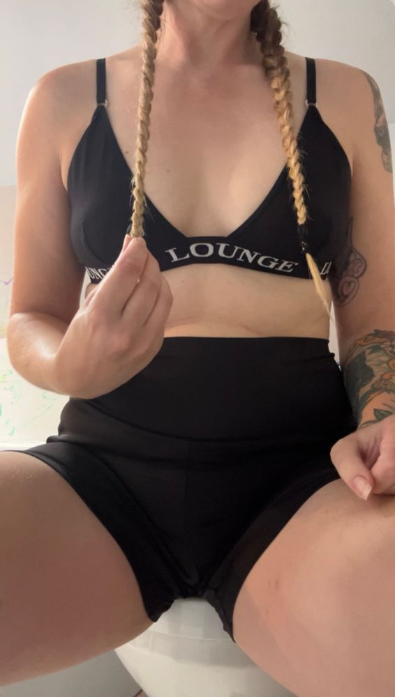 A woman’s torso from thighs to shoulders, she is wearing black shorts and a black bralette. Her blonde hair is tightly braided down past her nipples.
