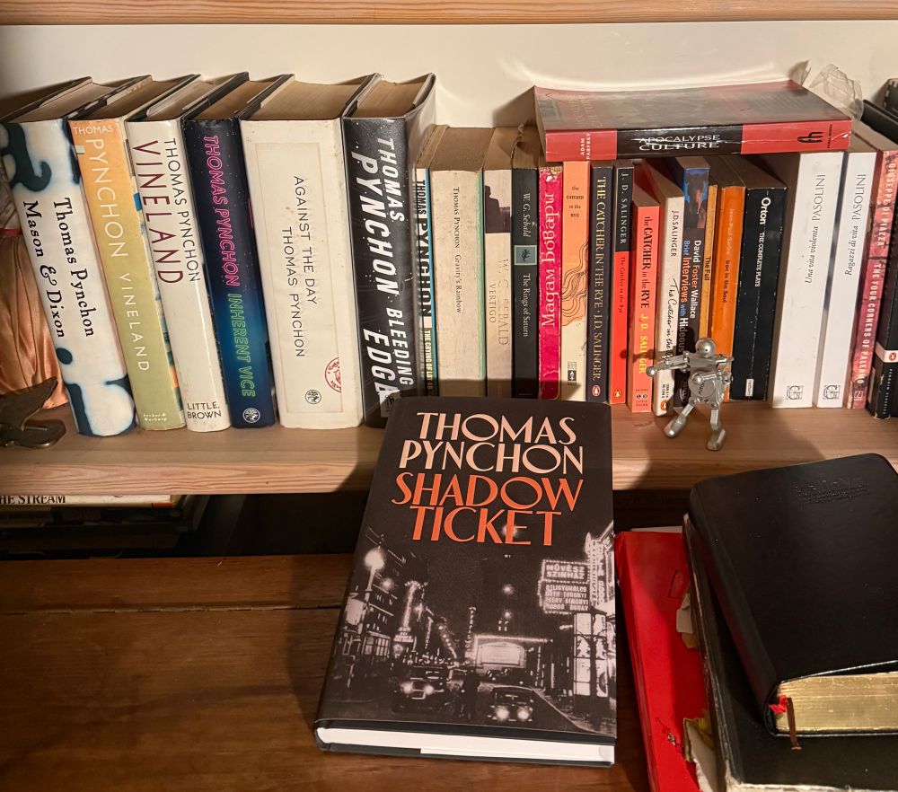 a bookshelf of Thomas Pynchon books with the latest release “Shadow Ticket” laid on the desk in front of it.