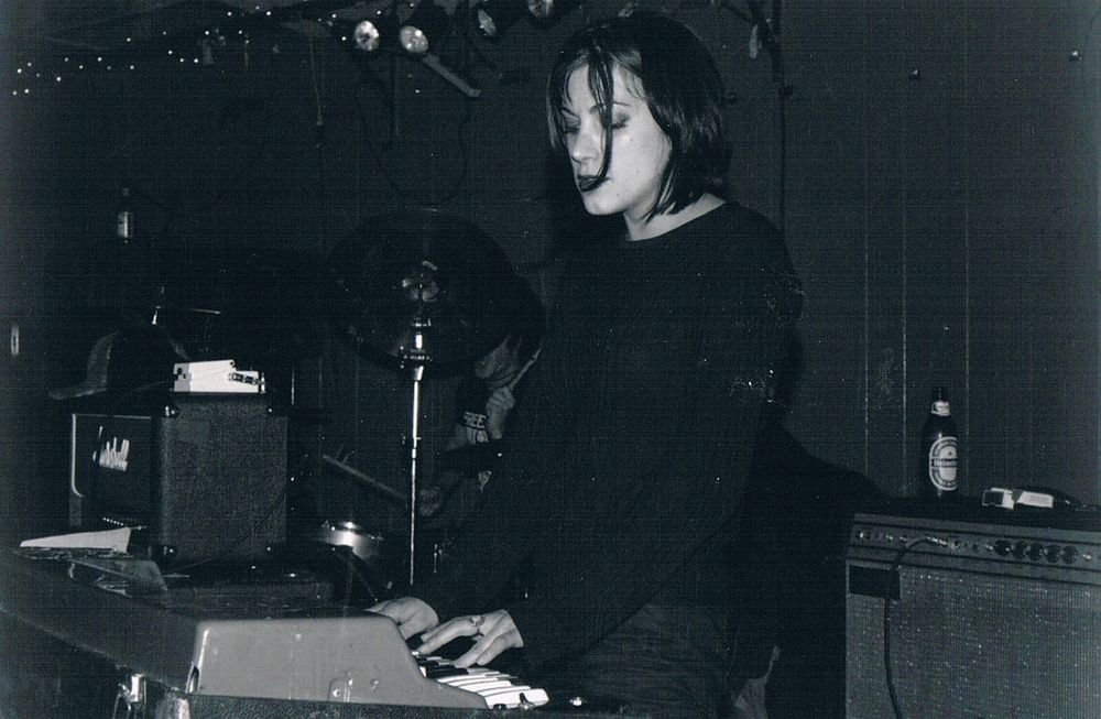 Black & white image. The keyboardist (leslie hardy) performs in a dimly lit venue, focused on playing a keyboard. She wears a black sweater, and in the background, but on the same stage you can see a drummer and amplifiers.