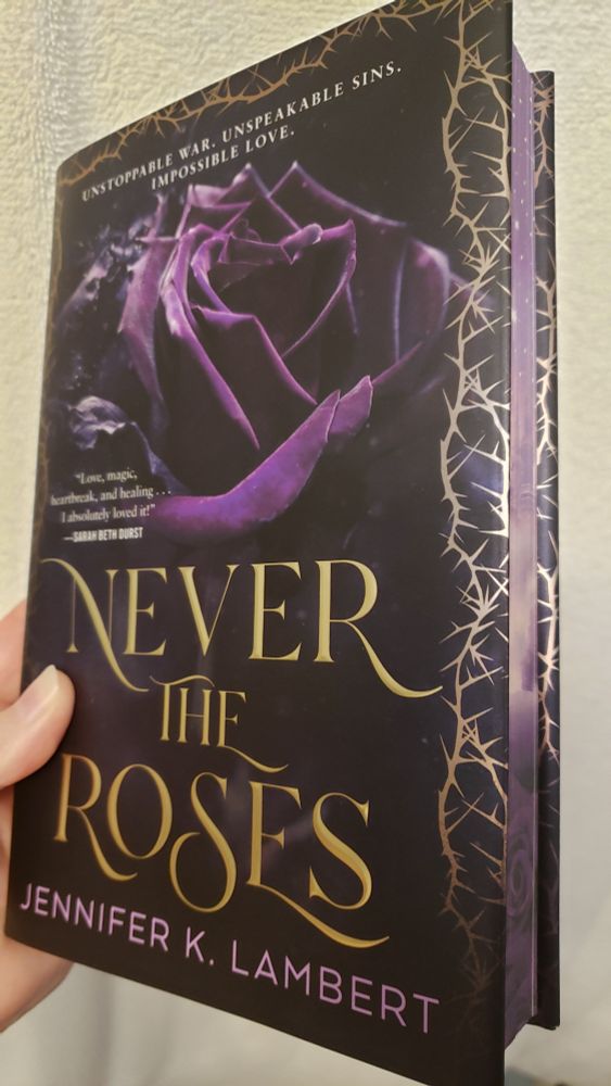 The fantasy novel "Never the Roses" by Jennifer Lambert, held at an angle to show not only the dark cover featuring a deep purple rose surrounded by thorns but also the purple sprayed edges.
