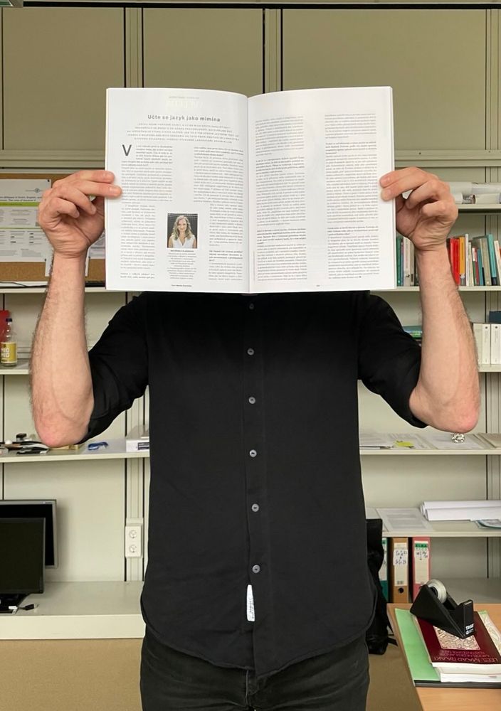 A person holding the April issue of the Czech bi-monthly magazine Heroine open in front of their face. The open pages show an interview with Kateřina Chládková on language acquisition. A bookcase is visible in the background.