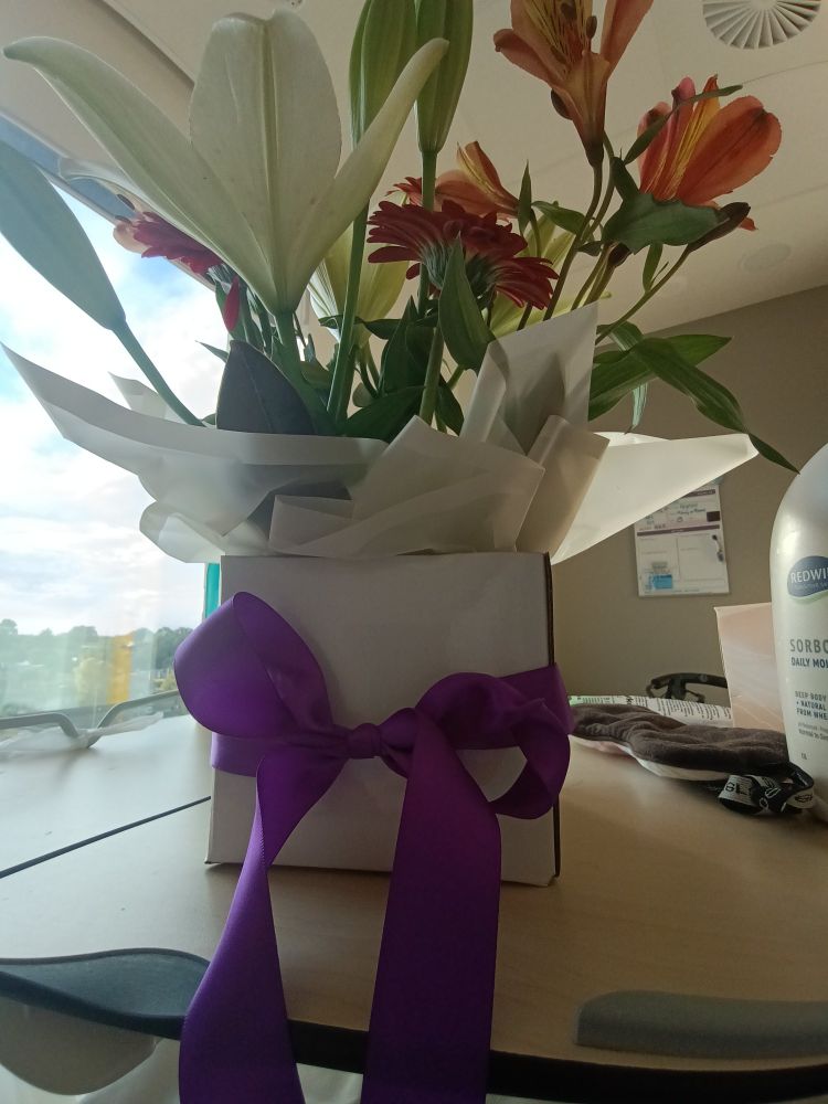 Box of flowers with purple ribbon. White lilies and red/ orange flowers. With generic hospital room and window in the background 