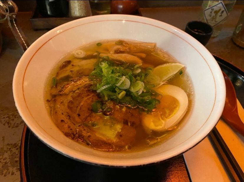 A bowl of ramen topped with chopped scallions, lime halves, half an egg, bamboo shoots, and pork belly.