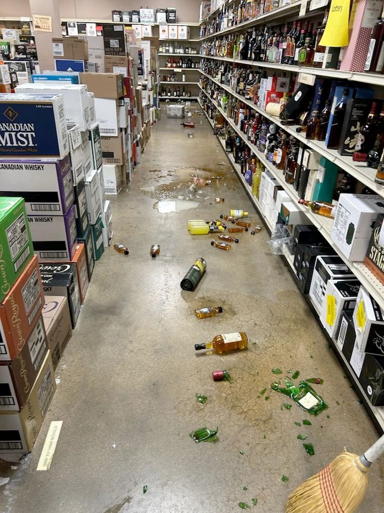 Liqor store aisle with many bottles strewed on the floor, some broken and areas of floor wet with alchool.