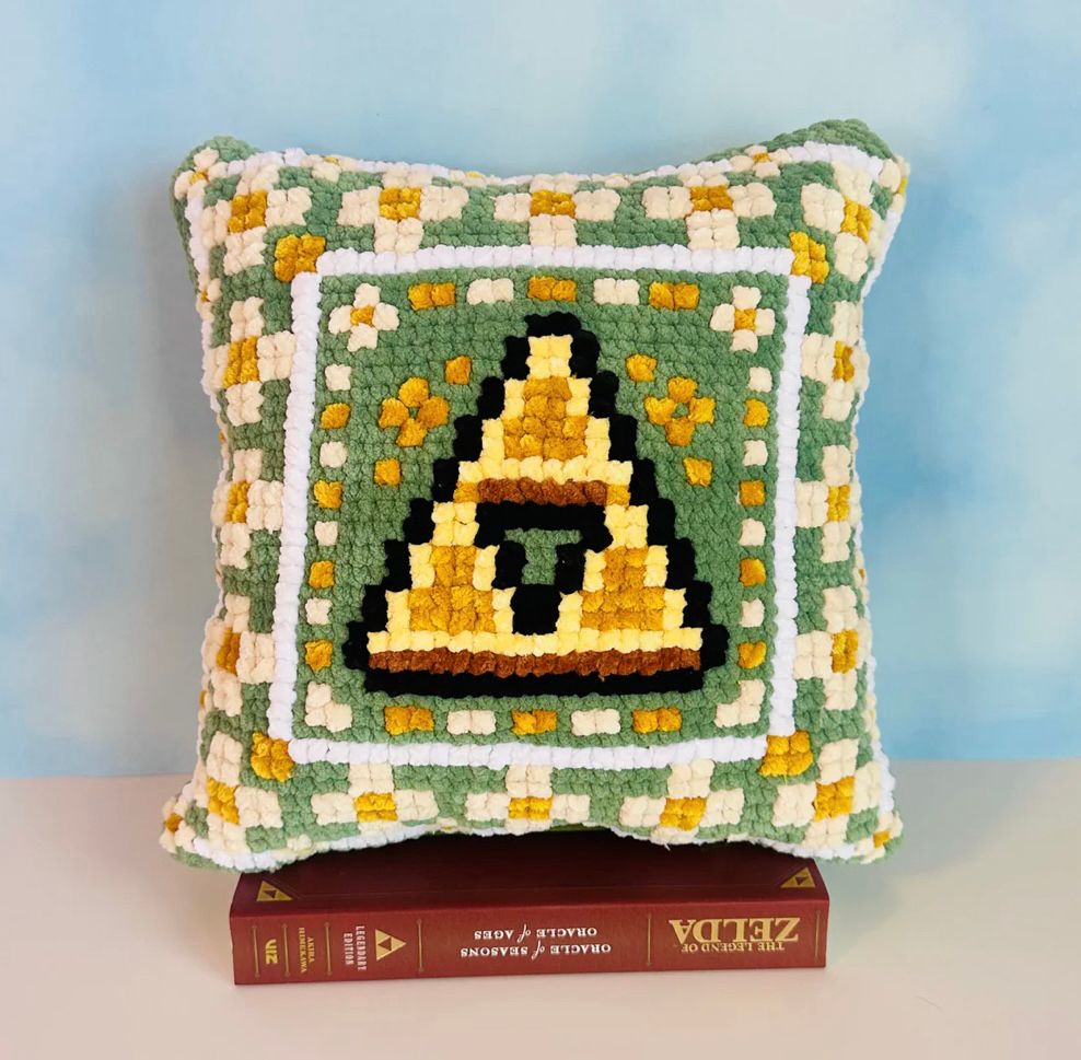 A handcrafted pillow featuring a crochet and cross-stitch design inspired by the Zelda video game series. The front showcases three red pixelated hearts with white borders, reminiscent of the game’s health icons, set against a light green background adorned with small daisy-like flowers.