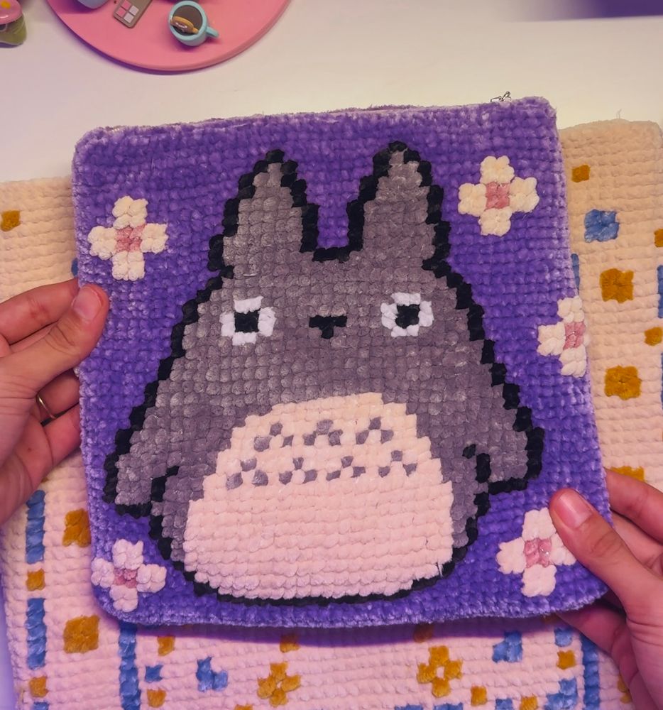 A handmade latch-hook or crochet project featuring a depiction of Totoro, a beloved animated character, on a purple background. The design is bordered by small pink and yellow flowers, adding a whimsical and colorful touch. The piece is held by two hands, showcasing its texture and detailing. The project rests on a patterned fabric with yellow and blue designs, creating a cozy, crafty atmosphere.