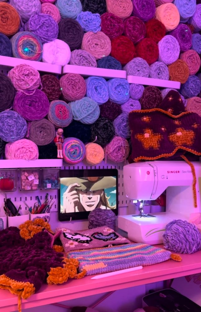 A vibrant, organized crafting space with a wall full of neatly arranged yarn in shades of pink, purple, and red. A sewing machine sits on the desk alongside crochet projects and tools, while a tablet displays a classic anime scene, creating a lively and creative atmosphere. The soft pink lighting enhances the warmth and charm of the workspace.
